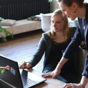 Two professional women discuss a project using a laptop in a modern office environment.